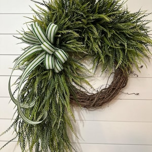 May include: A decorative wreath featuring a natural grapevine base, adorned with lush green faux foliage. A green and white striped bow adds a touch of elegance. The wreath is designed for seasonal home decor.
