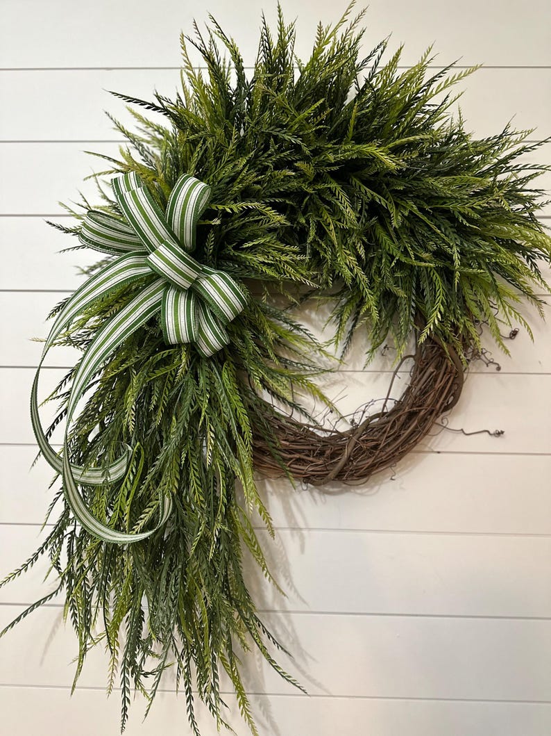 May include: A decorative wreath featuring a natural grapevine base, adorned with lush green faux foliage. A green and white striped bow adds a touch of elegance. The wreath is designed for seasonal home decor.