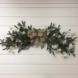 May include: A brown burlap bow tied around a garland of green olive branches and white flowers.