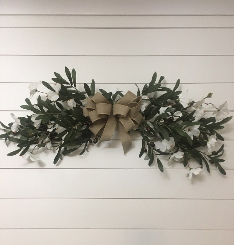 May include: A brown burlap bow tied around a garland of green olive branches and white flowers.