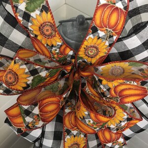 May include: A black and white buffalo plaid ribbon with a fall-themed ribbon featuring sunflowers, pumpkins, and leaves. The ribbon is tied into a bow.