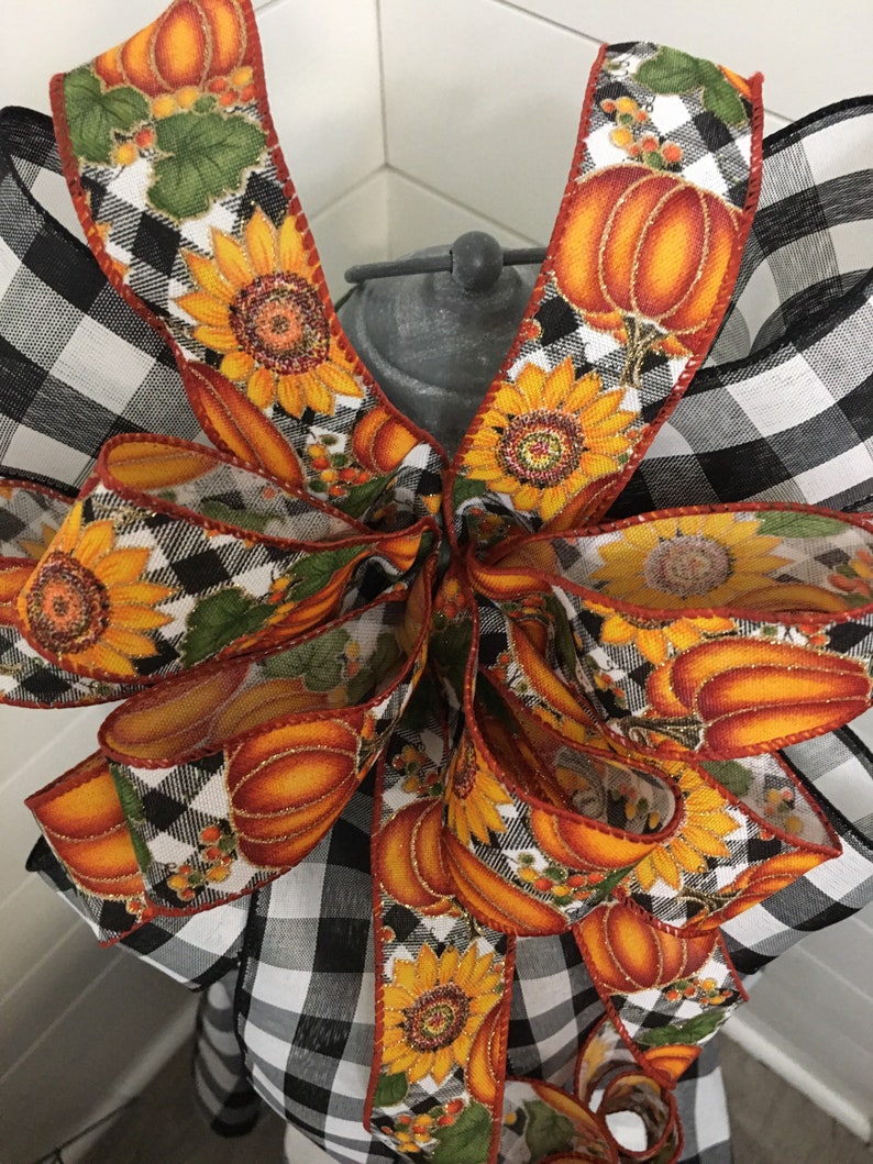 May include: A black and white buffalo plaid ribbon with a fall-themed ribbon featuring sunflowers, pumpkins, and leaves. The ribbon is tied into a bow.