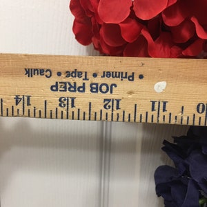 May include: A wooden ruler with black markings and the text "JOB PREP Primer Tape Caulk" is placed near red and navy blue artificial flowers. The ruler measures approximately 12 inches.