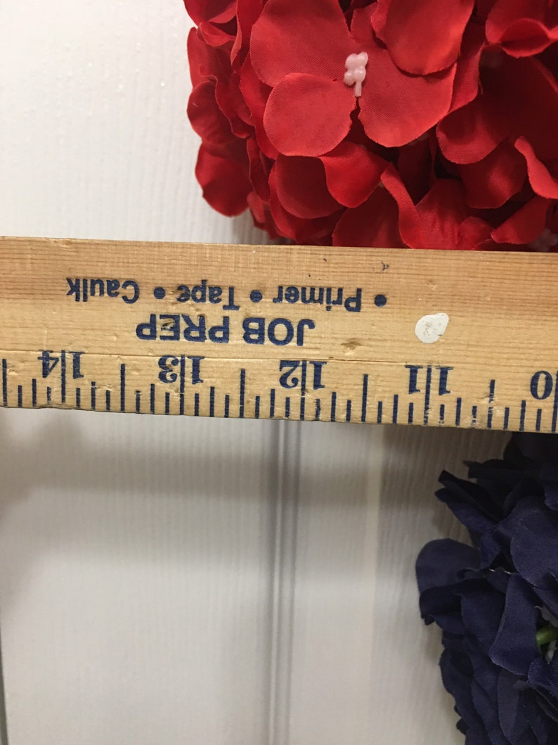 May include: A wooden ruler with black markings and the text "JOB PREP Primer Tape Caulk" is placed near red and navy blue artificial flowers. The ruler measures approximately 12 inches.