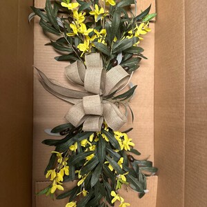 May include: A bouquet of yellow flowers with green leaves and brown berries tied with a burlap bow. The bouquet is in a cardboard box.