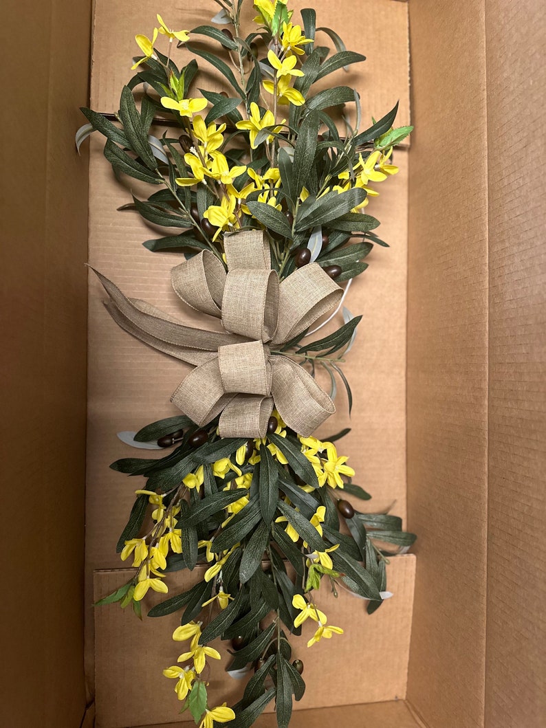 May include: A bouquet of yellow flowers with green leaves and brown berries tied with a burlap bow. The bouquet is in a cardboard box.