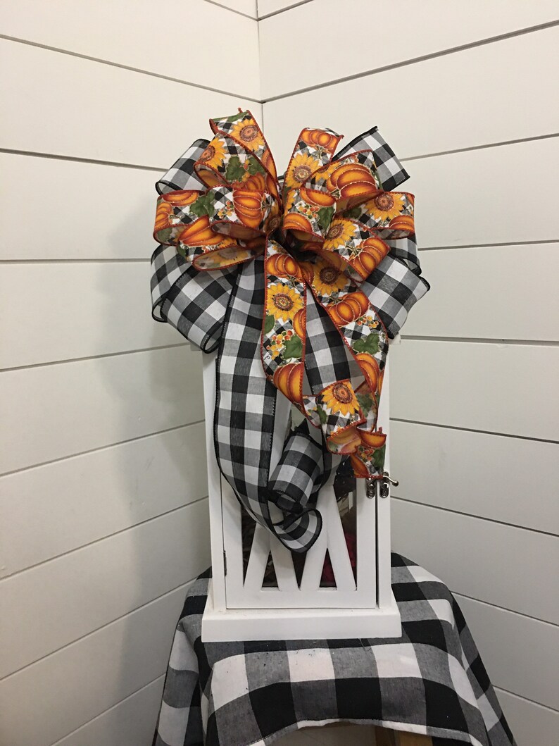 May include: A white lantern with a black and white checkered bow with a pumpkin and sunflower print ribbon. The lantern is sitting on a black and white checkered table cloth.