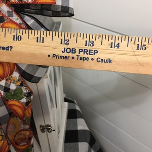May include: White lantern with a black and white checkered fabric base. The lantern is decorated with a fall-themed ribbon with pumpkins and sunflowers. The ribbon is tied with a bow. A ruler is placed next to the lantern, showing that it is approximately 13 inches tall. The ruler also has the text "JOB PREP" and "&bull; Primer &bull; Tape &bull; Caulk" printed on it.