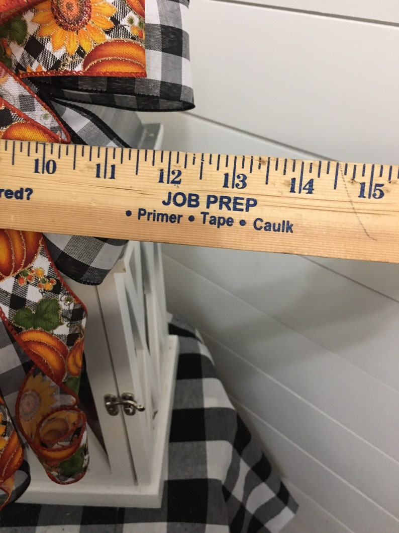 May include: White lantern with a black and white checkered fabric base. The lantern is decorated with a fall-themed ribbon with pumpkins and sunflowers. The ribbon is tied with a bow. A ruler is placed next to the lantern, showing that it is approximately 13 inches tall. The ruler also has the text "JOB PREP" and "&bull; Primer &bull; Tape &bull; Caulk" printed on it.