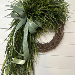 May include: A decorative wreath featuring a natural grapevine base, adorned with lush green faux foliage. A sage green velvet ribbon is tied in a bow and cascades down the side. The wreath is designed for door or wall decor.
