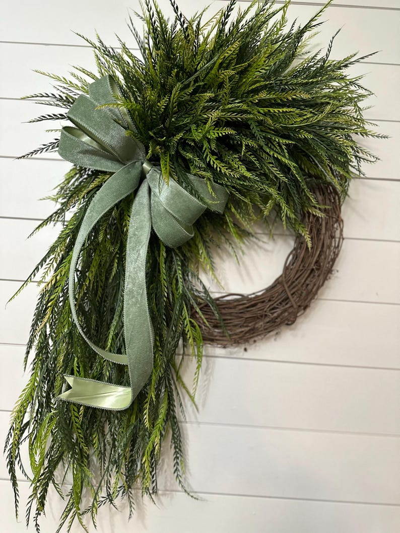 May include: A decorative wreath featuring a natural grapevine base, adorned with lush green faux foliage. A sage green velvet ribbon is tied in a bow and cascades down the side. The wreath is designed for door or wall decor.
