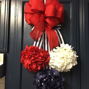 May include: A patriotic-themed door decoration featuring a large red bow with a striped ribbon, and three floral spheres in red, white, and navy blue. The arrangement is mounted on a dark blue door.