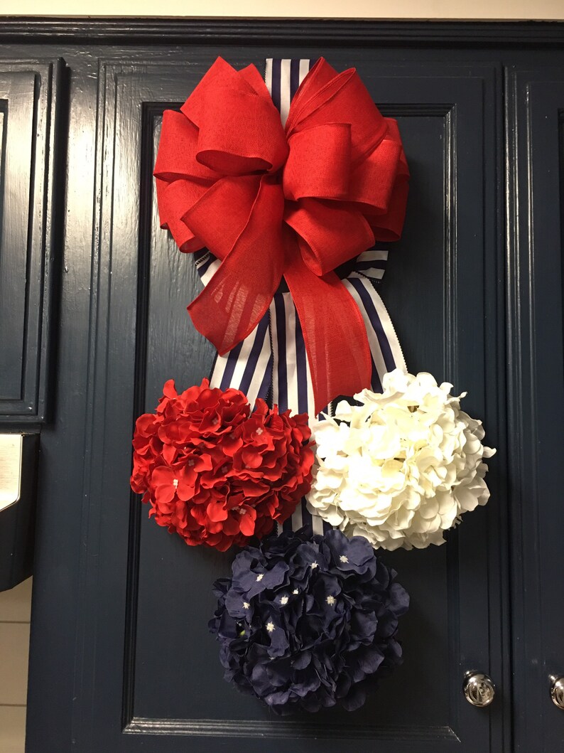 May include: A patriotic-themed door decoration featuring a large red bow with a striped ribbon, and three floral spheres in red, white, and navy blue. The arrangement is mounted on a dark blue door.