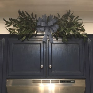 May include: A blue bow with a burlap texture is centered above two dark blue kitchen cabinets. The bow is surrounded by a garland of green leaves with small brown berries. A stainless steel range hood is below the cabinets.