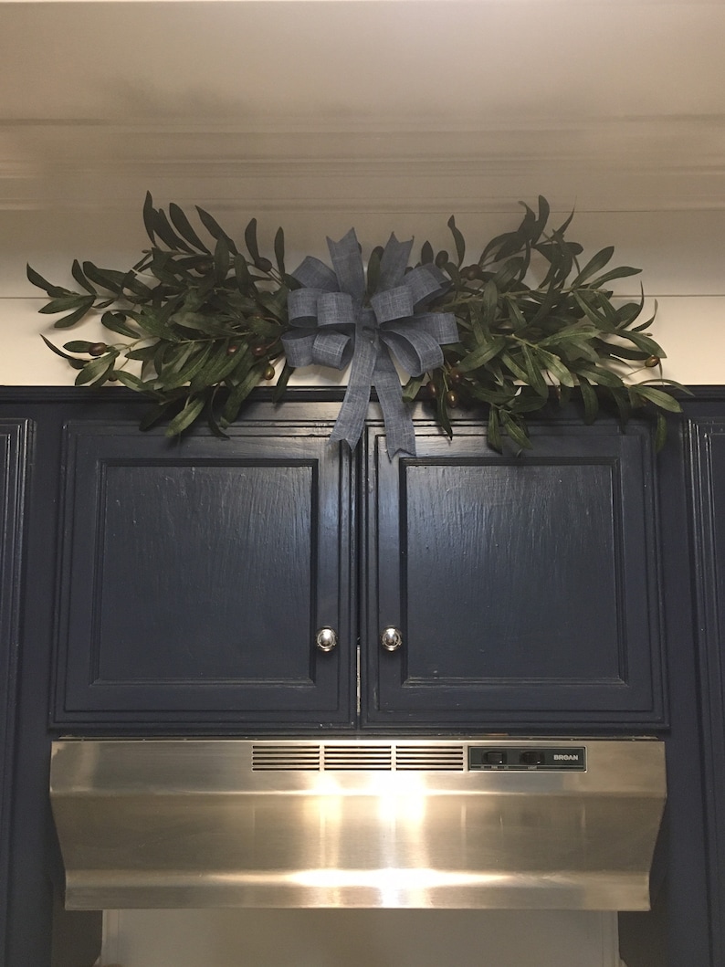 May include: A blue bow with a burlap texture is centered above two dark blue kitchen cabinets. The bow is surrounded by a garland of green leaves with small brown berries. A stainless steel range hood is below the cabinets.