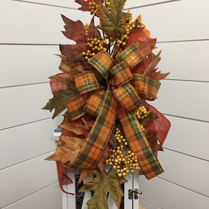 May include: A white lantern with a plaid bow and fall foliage, including orange, brown, and green leaves and yellow berries.