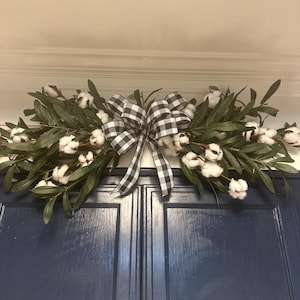 May include: A black and white checkered bow tied around two sprigs of greenery with white cotton balls. The greenery is draped over a set of dark blue cabinet doors.