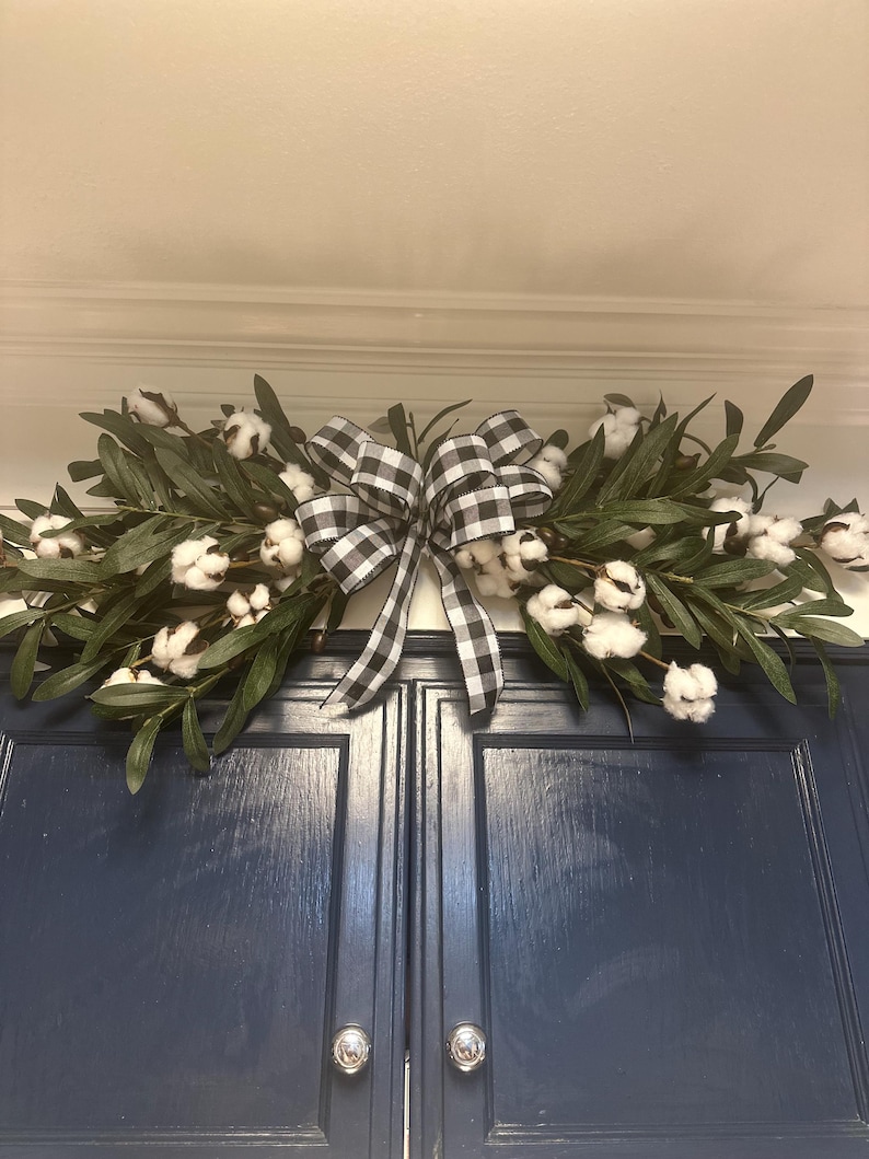 May include: A black and white checkered bow tied around two sprigs of greenery with white cotton balls. The greenery is draped over a set of dark blue cabinet doors.