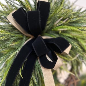 May include: Close-up of a decorative wreath featuring a large black velvet bow and tan burlap ribbon accents. The wreath is composed of green and tan faux foliage, creating a natural and elegant aesthetic. Perfect for seasonal home decor.