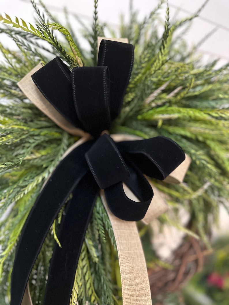 May include: Close-up of a decorative wreath featuring a large black velvet bow and tan burlap ribbon accents. The wreath is composed of green and tan faux foliage, creating a natural and elegant aesthetic. Perfect for seasonal home decor.