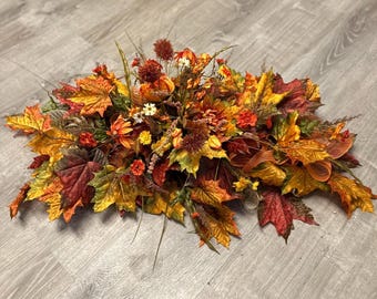 May include: An elongated floral arrangement featuring a mix of artificial autumn foliage, including maple leaves in shades of red, orange, and yellow, interspersed with small orange flowers, dried-look grasses, and textured brown seed heads.