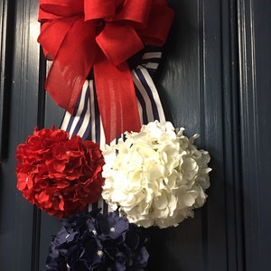 May include: A patriotic door decoration featuring a large red bow with a striped blue and white ribbon. Below the bow are three hydrangea flower balls in red, white, and navy blue. The arrangement is hanging on a dark blue door.