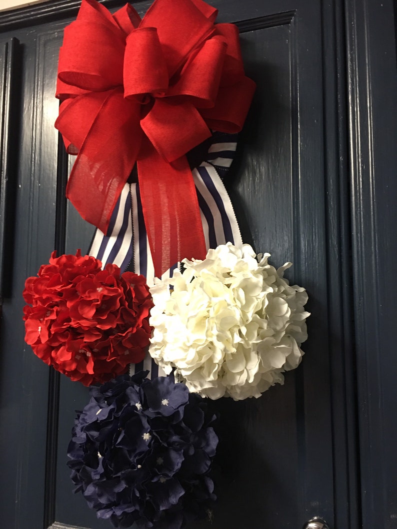 May include: A patriotic door decoration featuring a large red bow with a striped blue and white ribbon. Below the bow are three hydrangea flower balls in red, white, and navy blue. The arrangement is hanging on a dark blue door.