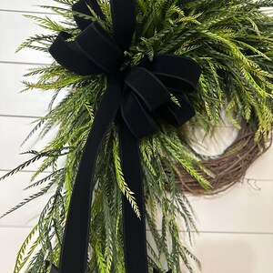 May include: A decorative wreath featuring a natural grapevine base, adorned with vibrant green foliage and a large black velvet bow. The bow's long, flowing tails add a touch of elegance to the design. Perfect for seasonal home decor.