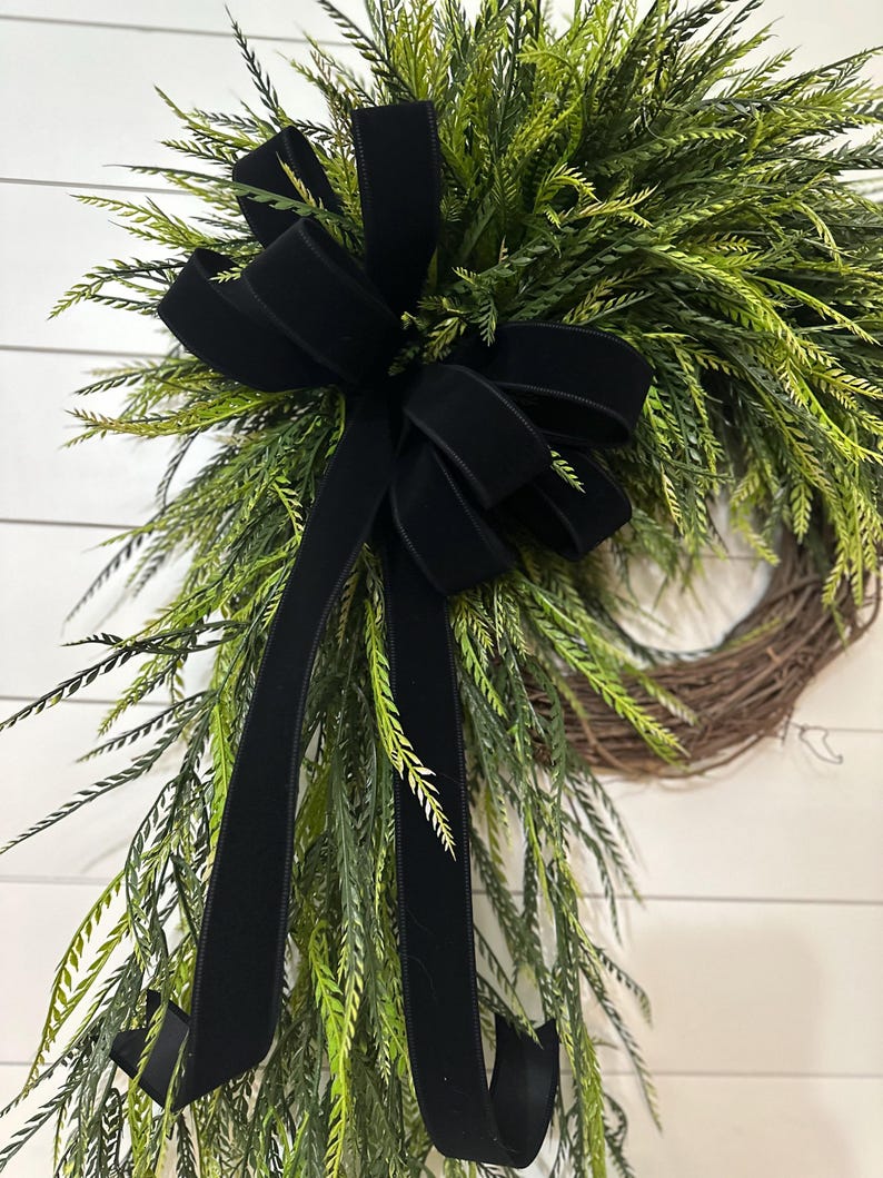May include: A decorative wreath featuring a natural grapevine base, adorned with vibrant green foliage and a large black velvet bow. The bow's long, flowing tails add a touch of elegance to the design. Perfect for seasonal home decor.