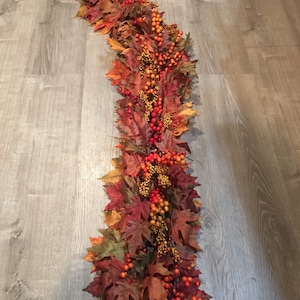 May include: A fall-themed garland featuring faux maple leaves in shades of red, orange, and brown. The garland is adorned with clusters of orange and red berries, creating a festive and autumnal decorative piece.