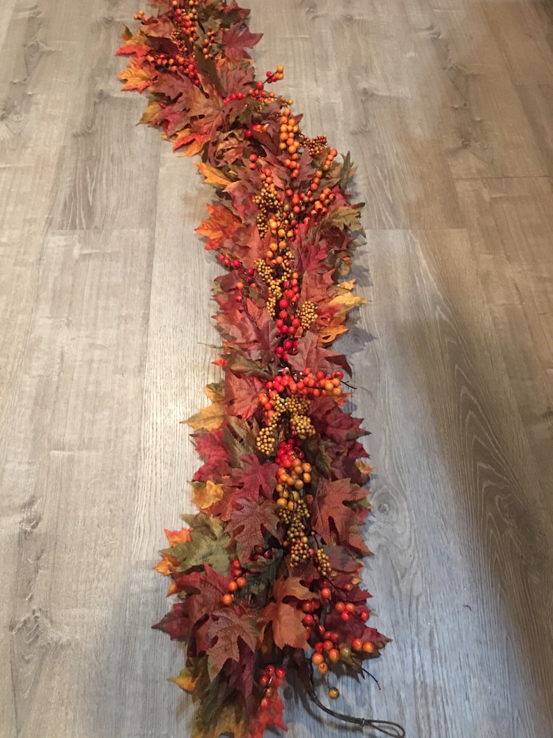 May include: A fall-themed garland featuring faux maple leaves in shades of red, orange, and brown. The garland is adorned with clusters of orange and red berries, creating a festive and autumnal decorative piece.