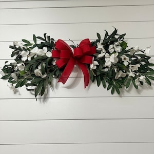 May include: A white and green floral swag with a large red bow. The swag is hanging on a white wall.