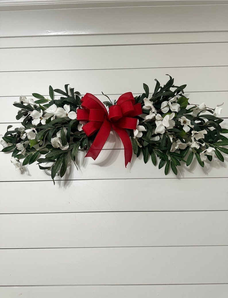 May include: A white and green floral swag with a large red bow. The swag is hanging on a white wall.