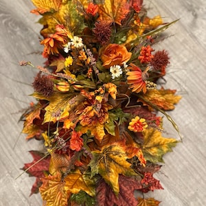 May include: A fall-themed floral arrangement featuring a mix of artificial maple leaves in shades of red, orange, and yellow. The arrangement includes orange and yellow flowers, and brown accents. The centerpiece is designed for table decor.