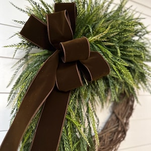 May include: A decorative wreath featuring a brown velvet bow and lush green foliage. The wreath is made of woven twigs, creating a natural and rustic aesthetic. The bow adds a touch of elegance to the overall design, suitable for home decor.