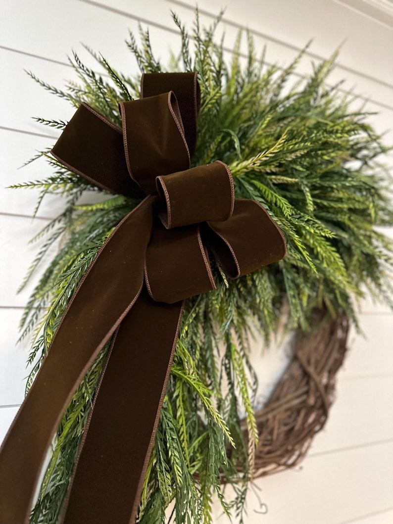 May include: A decorative wreath featuring a brown velvet bow and lush green foliage. The wreath is made of woven twigs, creating a natural and rustic aesthetic. The bow adds a touch of elegance to the overall design, suitable for home decor.