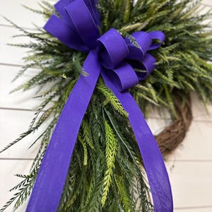 May include: A decorative wreath featuring a vibrant purple bow and flowing ribbons. The wreath is adorned with green and tan faux foliage, creating a natural and inviting aesthetic. The bow is the focal point, adding a pop of color and elegance to the design.