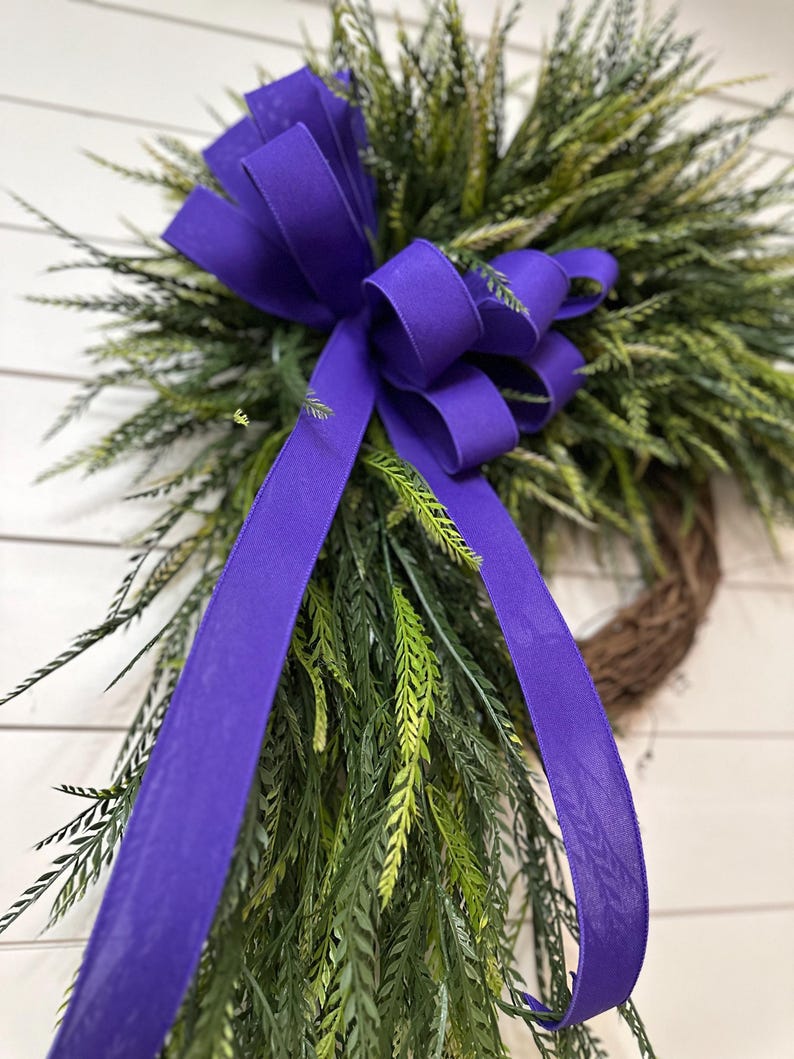 May include: A decorative wreath featuring a vibrant purple bow and flowing ribbons. The wreath is adorned with green and tan faux foliage, creating a natural and inviting aesthetic. The bow is the focal point, adding a pop of color and elegance to the design.