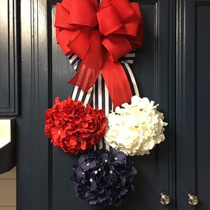 May include: A patriotic-themed decoration featuring a large red bow with a striped ribbon, and three floral spheres in red, white, and navy blue. The arrangement is mounted on a dark blue cabinet door.