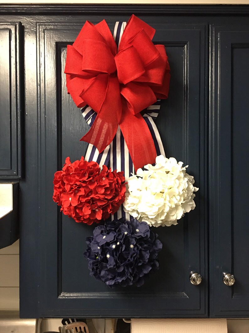 May include: A patriotic-themed decoration featuring a large red bow with a striped ribbon, and three floral spheres in red, white, and navy blue. The arrangement is mounted on a dark blue cabinet door.
