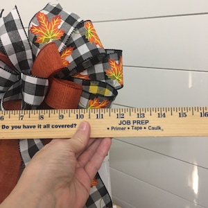 May include: A black and white checkered ribbon with orange and yellow maple leaf designs is tied into a bow. The bow is being held by a hand and a ruler is placed on top of the hand. The ruler reads "Do you have it all covered?" and "JOB PREP &bull; Primer &bull; Tape &bull; Caulk."