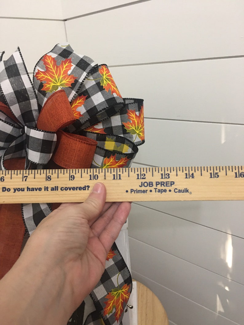 May include: A black and white checkered ribbon with orange and yellow maple leaf designs is tied into a bow. The bow is being held by a hand and a ruler is placed on top of the hand. The ruler reads "Do you have it all covered?" and "JOB PREP &bull; Primer &bull; Tape &bull; Caulk."