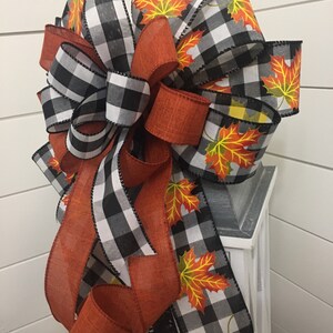May include: A black and white buffalo plaid ribbon with orange ribbon and orange and yellow maple leaf print ribbon. The ribbon is tied into a bow and is attached to a white lantern.