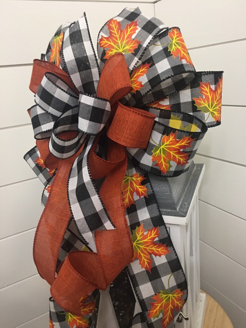 May include: A black and white buffalo plaid ribbon with orange ribbon and orange and yellow maple leaf print ribbon. The ribbon is tied into a bow and is attached to a white lantern.