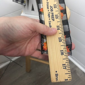 May include: A wooden ruler with black and white checkered ribbon with orange and yellow fall leaves printed on it. The ruler has markings in inches from 10 to 18. The ruler has the text "JOB PREP" and "Primer - Tape - Caulking" printed on it.