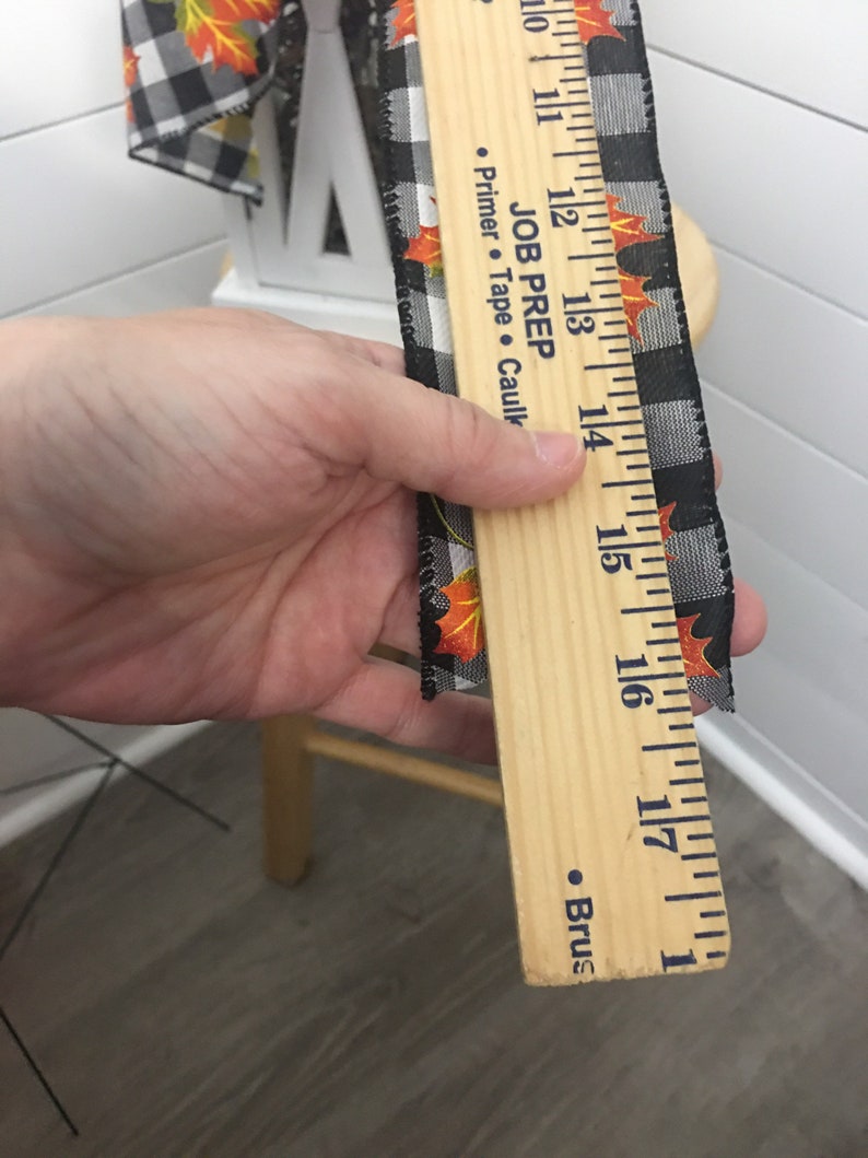 May include: A wooden ruler with black and white checkered ribbon with orange and yellow fall leaves printed on it. The ruler has markings in inches from 10 to 18. The ruler has the text "JOB PREP" and "Primer - Tape - Caulking" printed on it.