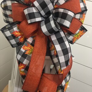 May include: A large bow made with black and white buffalo check ribbon and orange ribbon with a fall leaf print. The bow is tied with a loop and tails.
