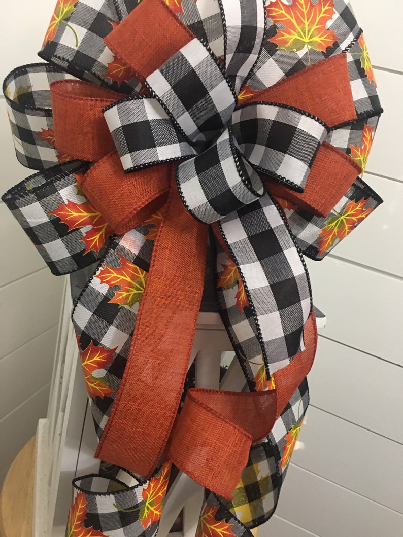 May include: A large bow made with black and white buffalo check ribbon and orange ribbon with a fall leaf print. The bow is tied with a loop and tails.