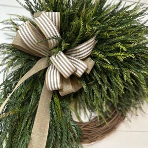 May include: A decorative wreath featuring lush green foliage and a brown and white striped bow. The wreath is made of natural-looking greenery and is perfect for adding a touch of nature to any home decor.
