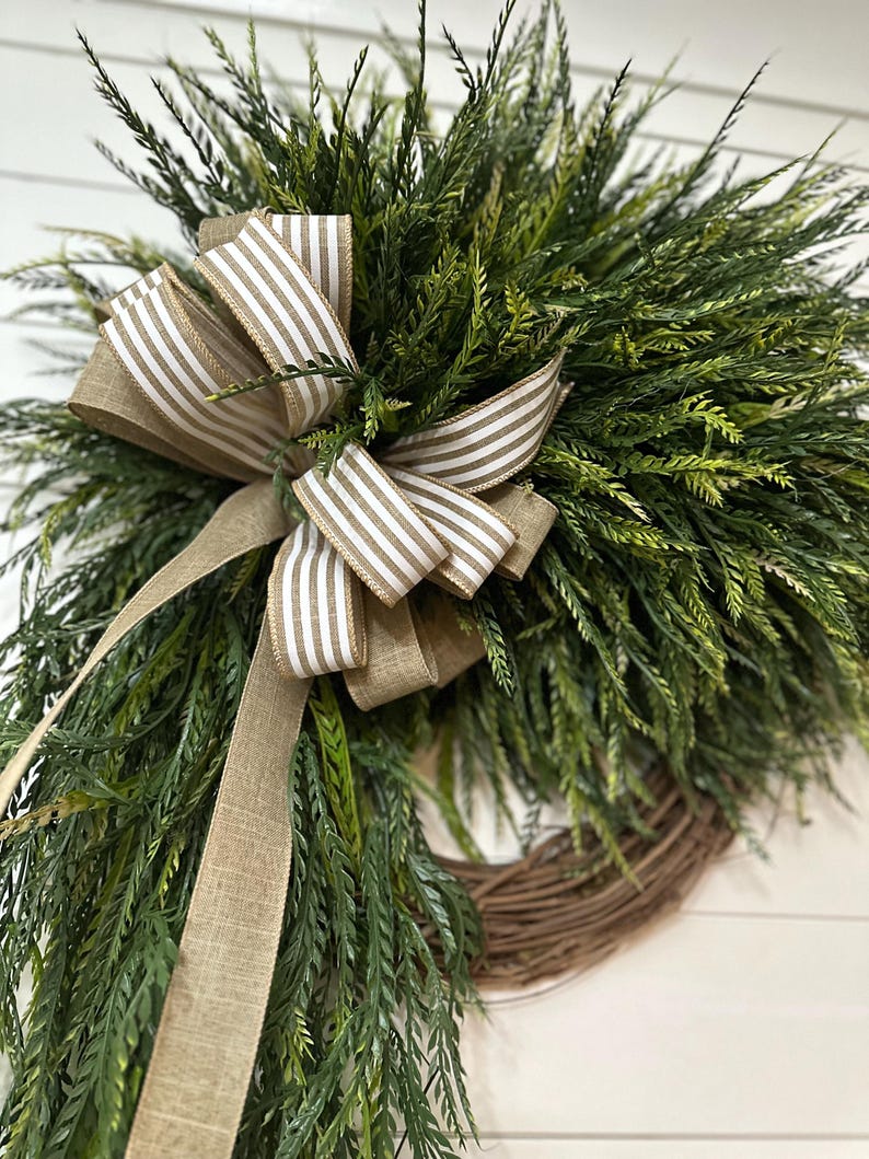 May include: A decorative wreath featuring lush green foliage and a brown and white striped bow. The wreath is made of natural-looking greenery and is perfect for adding a touch of nature to any home decor.